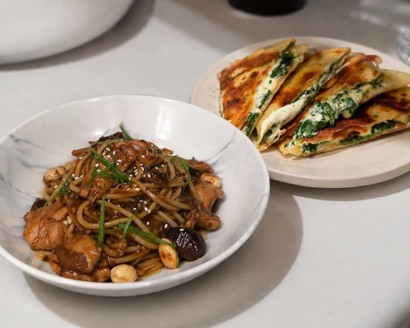 A bowl of noodles with sesame and greens beside toasted, filled flatbreads at BRGR.BEAN